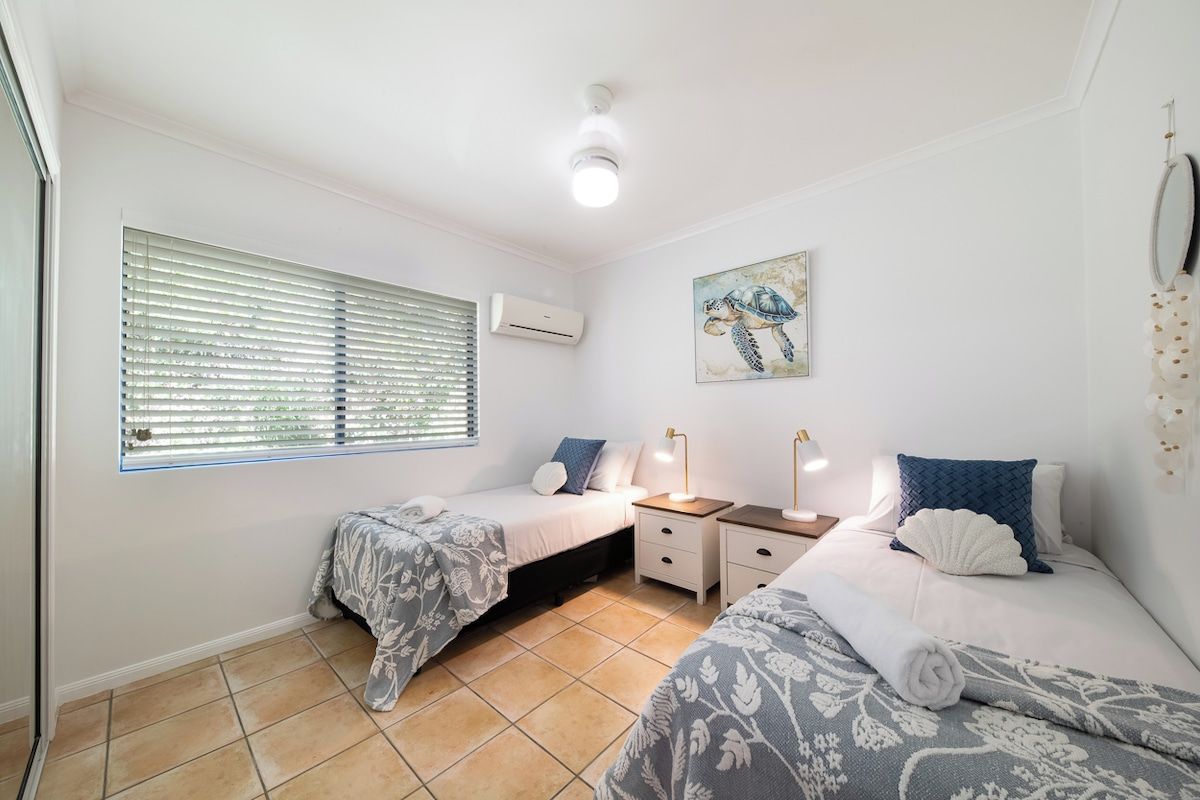 Une chambre avec deux lits, une fenêtre avec des stores, un appareil de climatisation, une table de chevet avec des lampes et un miroir fixé au mur.
