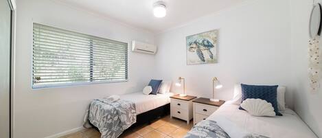 Une chambre avec deux lits, une fenêtre avec des stores, un appareil de climatisation, une table de chevet avec des lampes et un miroir fixé au mur.