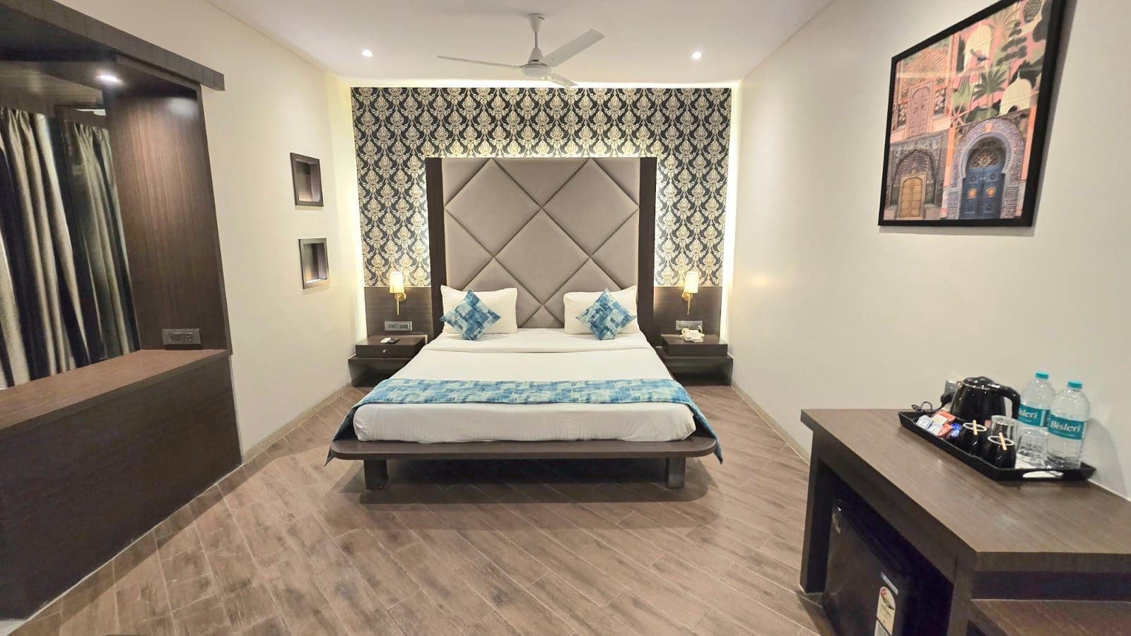 A hotel room with a bed, bedside tables, a desk, a chair, a ceiling fan, and a wall-mounted artwork.