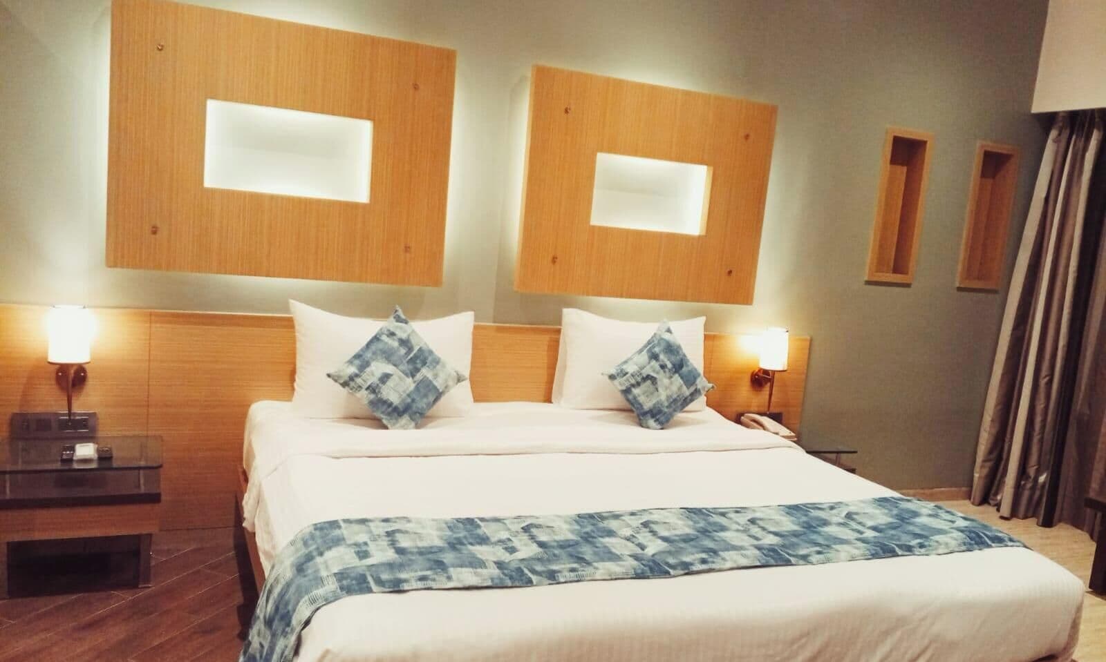 Habitación de hotel con una cama grande, dos mesitas de noche, cabecera con iluminación integrada y una ventana con cortinas.