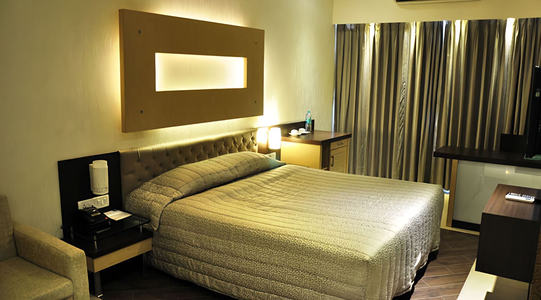 A hotel room with a bed, bedside table, lamp, and a view of curtains.
