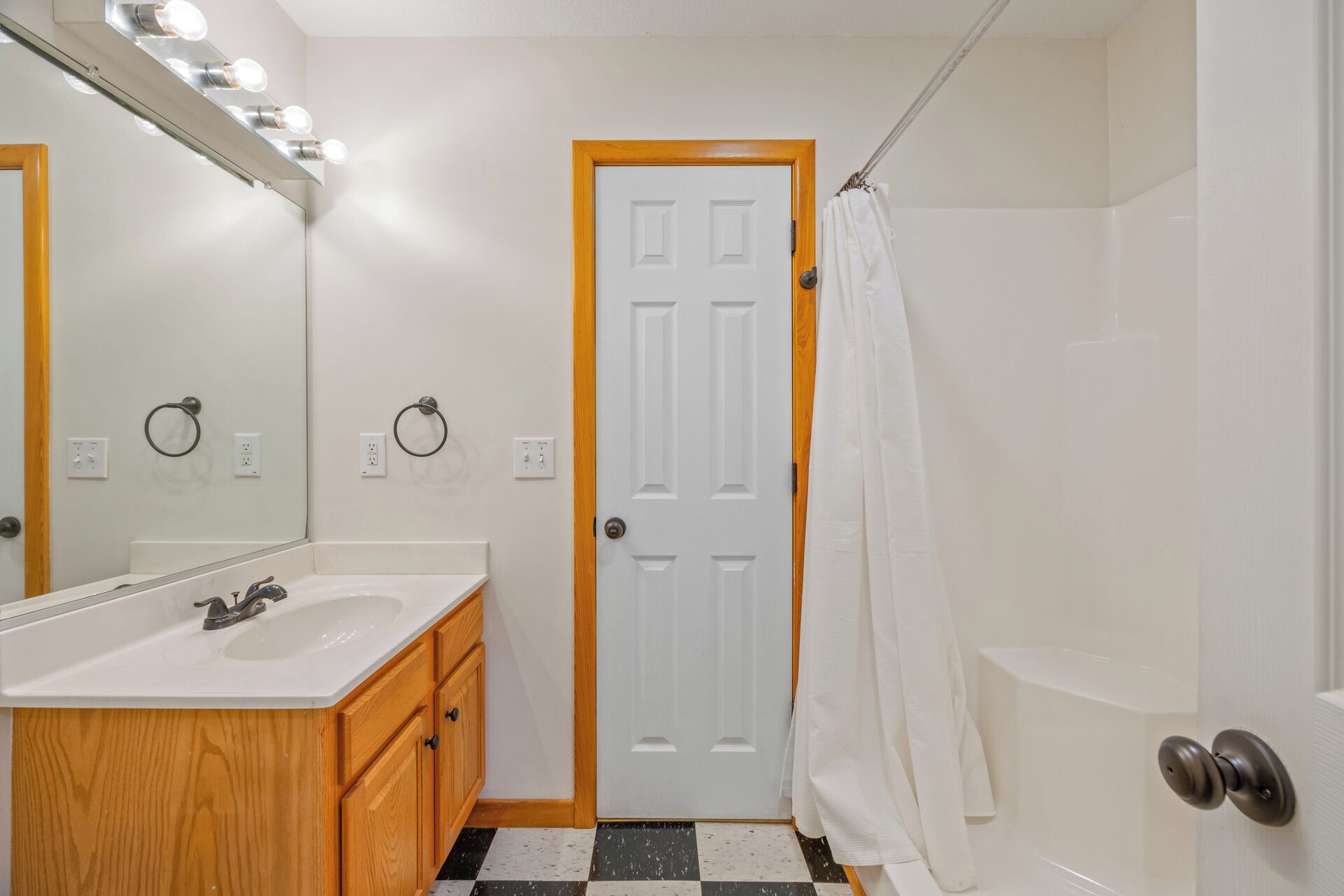 A bathroom with a white sink, wooden vanity, a mirror with lights, a white door, and a shower curtain.