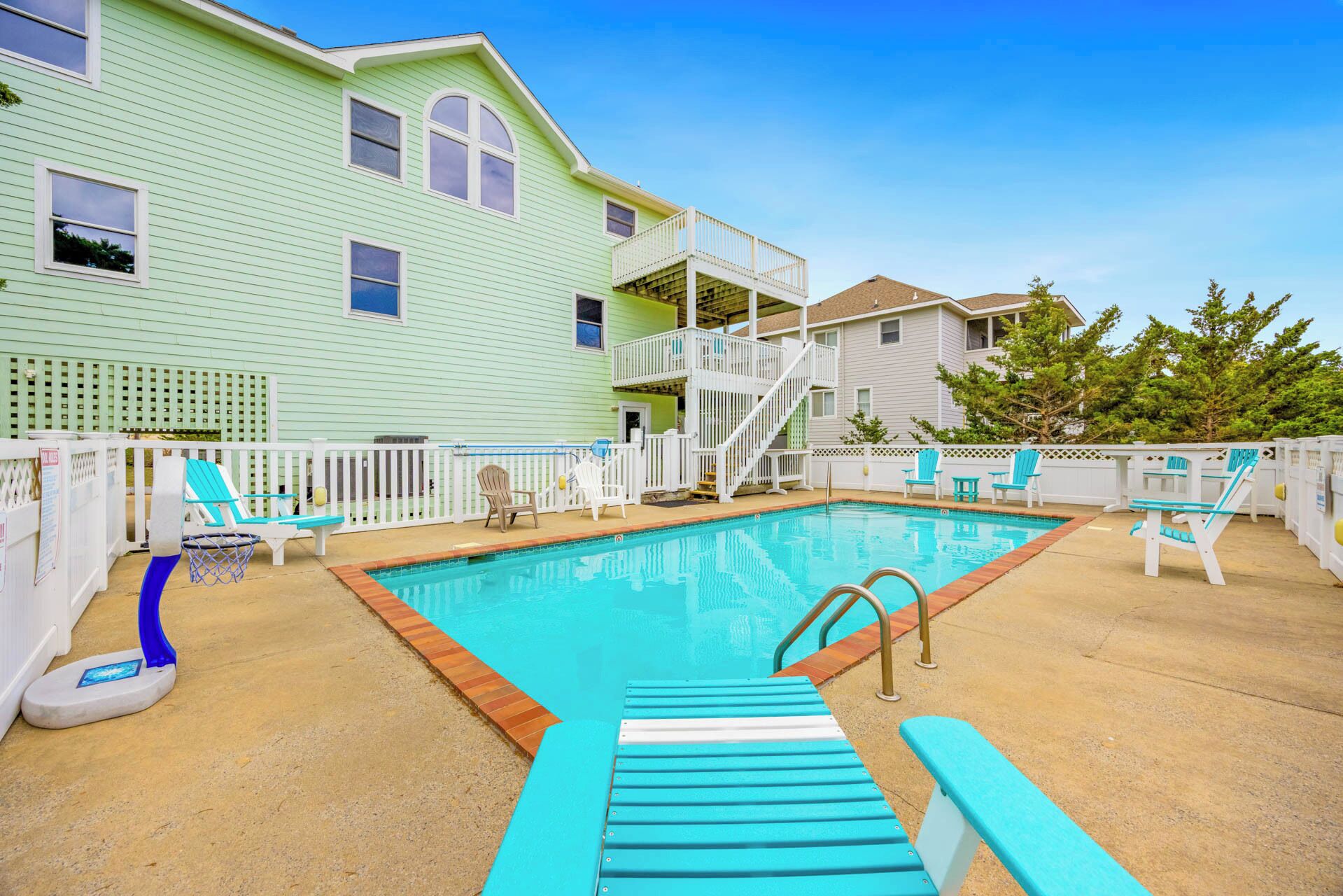 A swimming pool with a wooden deck, lounge chairs, and a white fence.