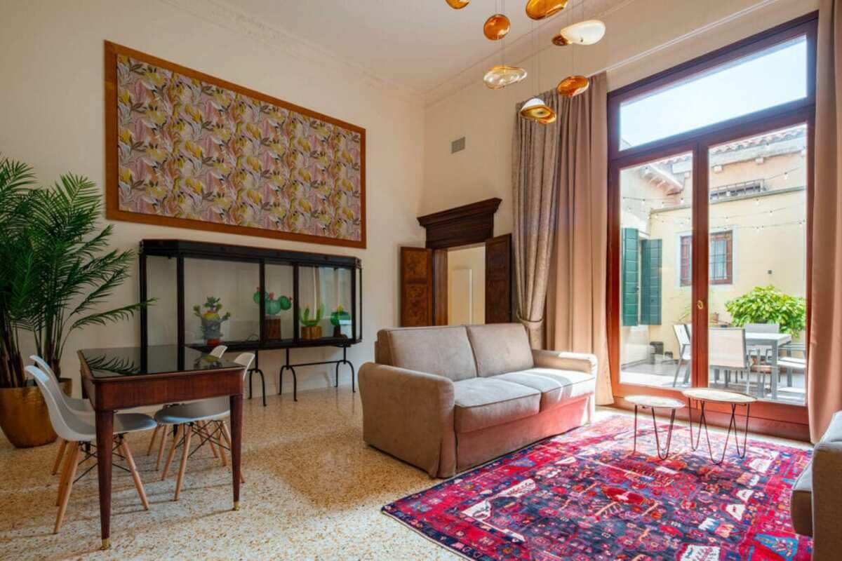 Ein Wohnzimmer mit einer Couch, einem Couchtisch, einem Esstisch mit Stühlen, einem großen Fenster mit Vorhängen und einem farbenfrohen Teppich.