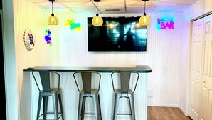 Zona de bar con televisor, tres taburetes y letreros de neón.
