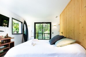 Ein Schlafzimmer mit einem Bett, einem hölzernen Kopfteil, einem Fernseher, einem Bücherregal und einem Fenster mit Blick auf Grünfläche.