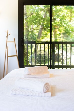 Une chambre à coucher avec un lit, des serviettes blanches, une échelle en bois et une vue sur la verdure, visible à travers la porte d’un balcon.