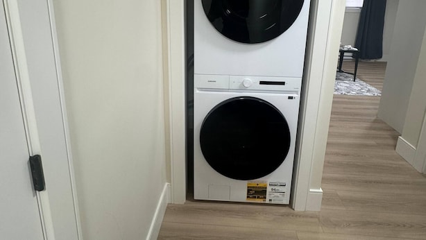 Un lavadero con lavadora y secadora de carga frontal, suelo de madera y un pasillo que conduce a otra habitación.