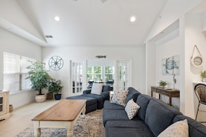 A spacious living room with a dark grey sofa, a wooden coffee table, and a large window with blinds. - Shipyard Golf Master - Spacious 3 bedroom Beach, Pool, Tennis & Pickleball ! (Hilton Head Island)