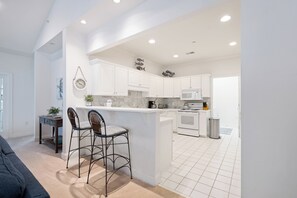 A modern kitchen with white cabinets, a central island, and a dining area with two bar stools. - Shipyard Golf Master - Spacious 3 bedroom Beach, Pool, Tennis & Pickleball ! (Hilton Head Island)