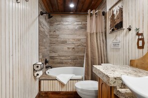 A bathroom with a wooden wall, a bathtub, a toilet, and a sink with a marble countertop. - Haskell Cabin 2 Bedroom Cabin (Grand Lake)