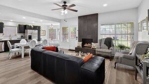 A modern living room with a black leather sofa, a dining area with white chairs, a kitchen with white cabinets, and a fireplace. - Central Frisco Family Getaway 3 Bedroom Home (Frisco)