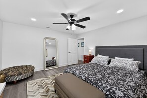 Une chambre à coucher comprenant un lit, un ventilateur de plafond, une table de chevet, un miroir et un panier pour animal de compagnie.
