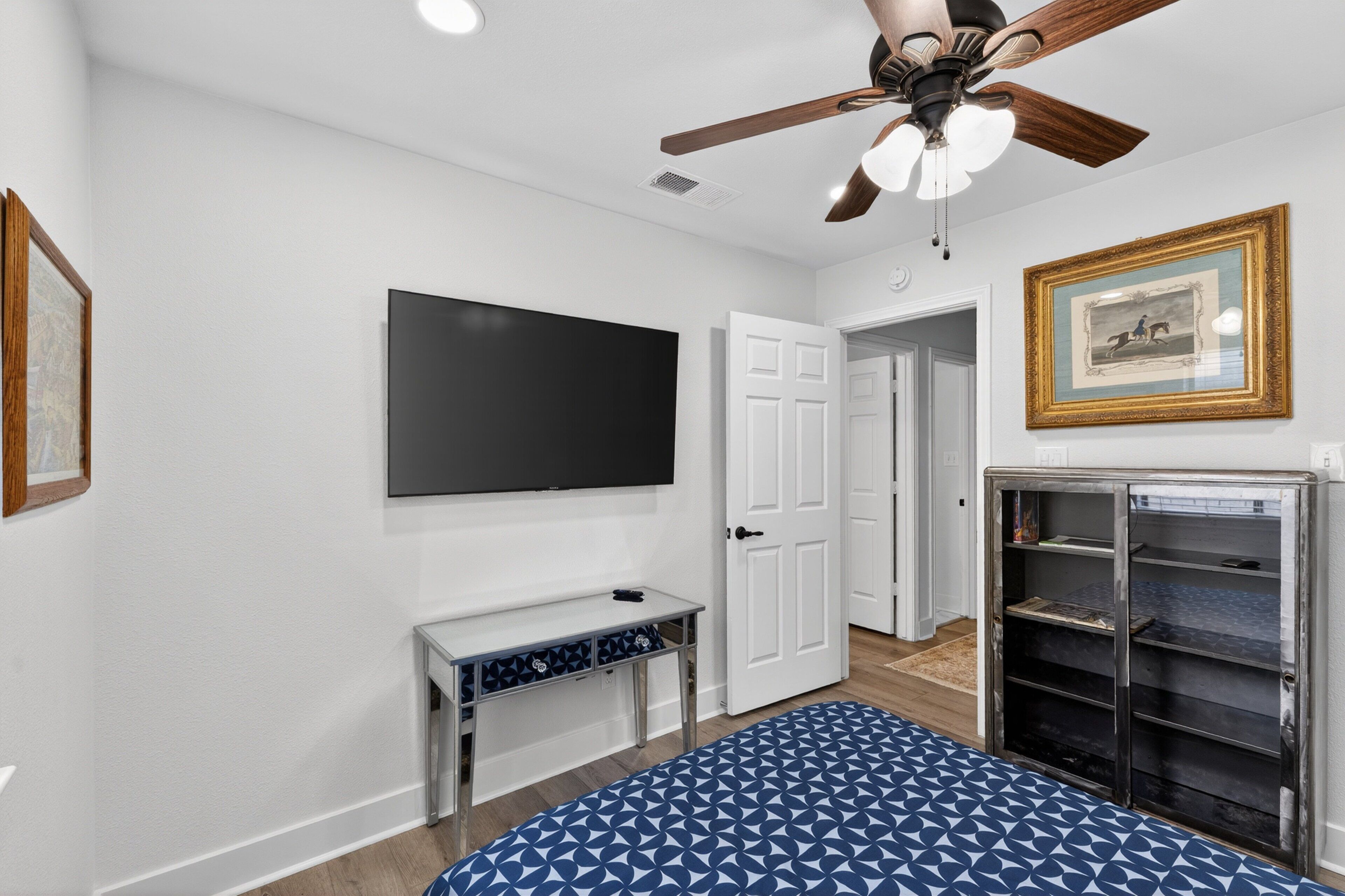 A bedroom with a ceiling fan, a TV mounted on the wall, a bed with a patterned bedspread, a white door, and a bookshelf.