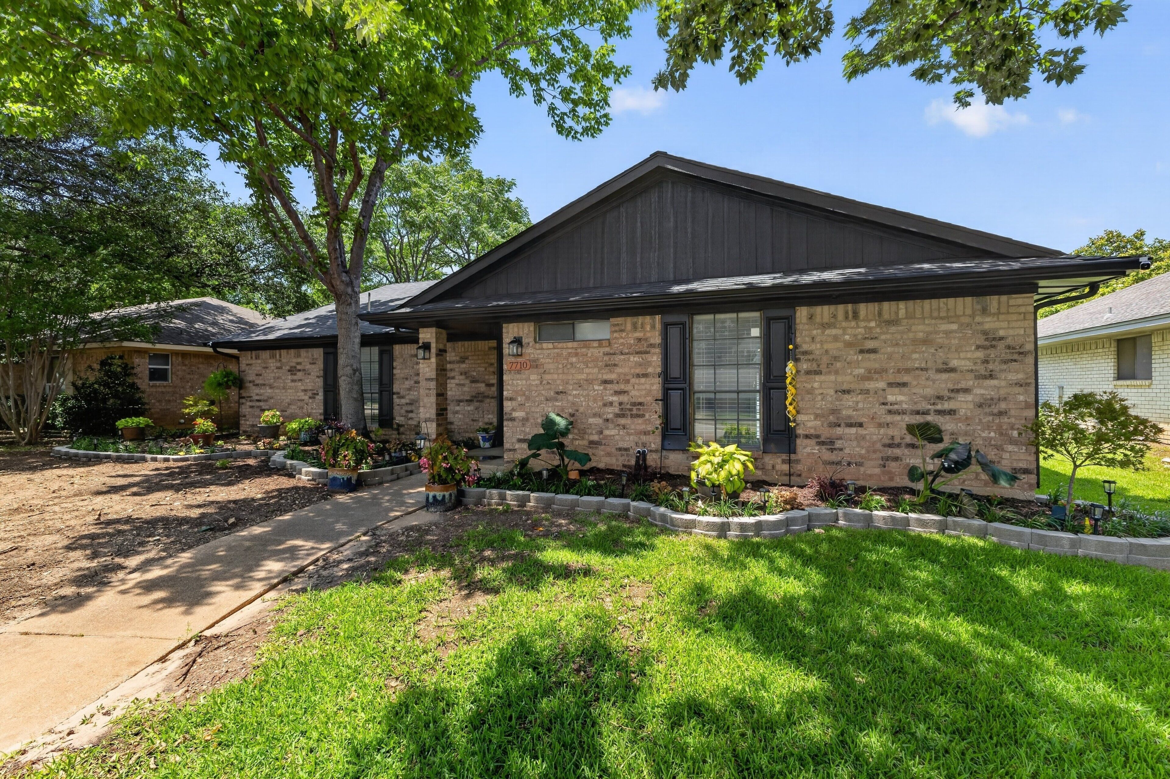 A single-story brick house with a dark roof, a front yard with a lawn and potted plants, and a clear blue sky.