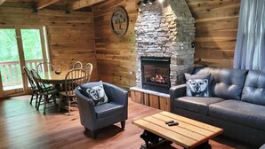 Interior de una cabaña de madera con chimenea de piedra, una mesa de comedor con sillas, un sofá y una mesa auxiliar.