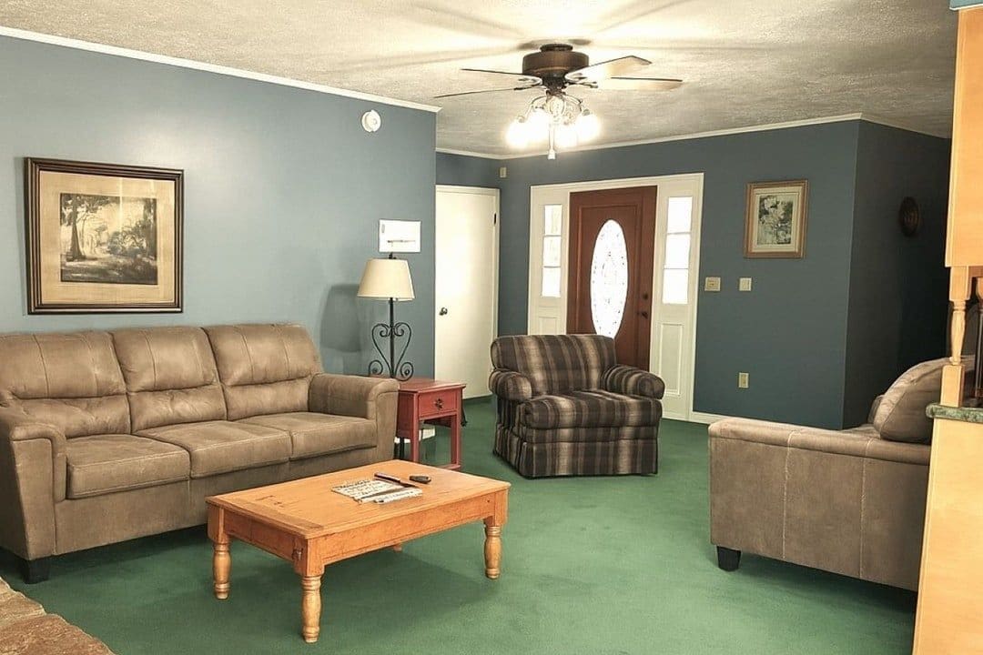 Una sala de estar con ventilador de techo, un sofá de cuero marrón, un sillón a cuadros y una mesa de centro de madera.