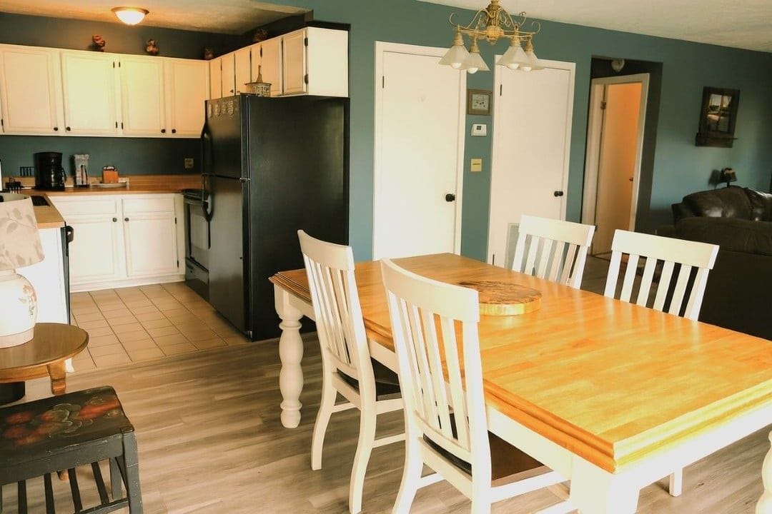 A compact kitchen with white cabinetry, a black refrigerator, and a wooden dining table with white chairs.