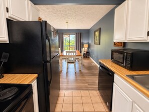 A kitchen with a black refrigerator, a microwave, and a stove. There is a dining table with chairs and a lamp. The walls are painted blue, and there is a window with curtains. - Hillbilly Hilton 2 Bedroom Chalet (Pigeon Forge)
