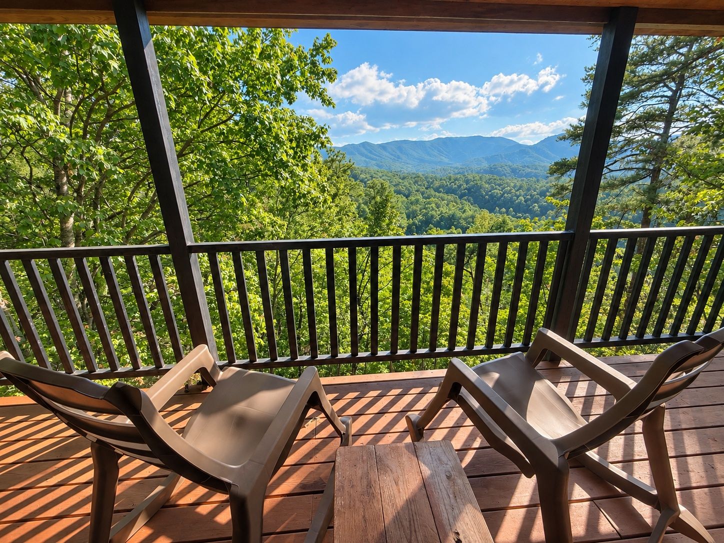 A wooden deck with two lounge chairs and a table, overlooking a forested mountain range.