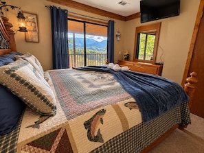 Un dormitorio con una cama grande, vistas a las montañas desde un balcón, una televisión y un cabecero de madera.