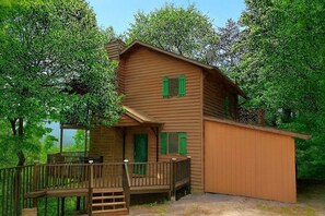 Eine zweistöckige Holzhütte mit Veranda und kleiner Terrasse, umgeben von Bäumen.