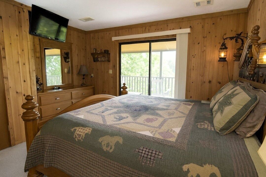 A bedroom with a bed, a dresser, a television mounted on the wall, and a balcony with a railing.