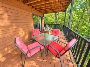 Una terraza techada al aire libre con una mesa de vidrio rodeada por cuatro sillas con cojines rojos, baranda de madera y vista a árboles.