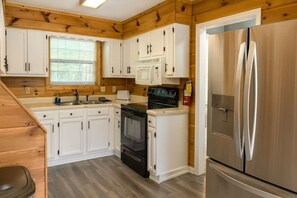 Cocina compacta con alacenas blancas, estufa negra, refrigerador de acero inoxidable y ventana con persianas.
