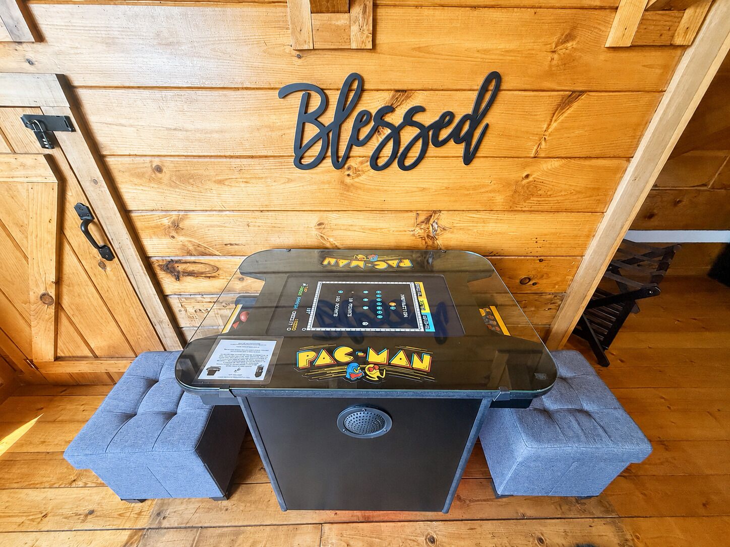 Ein Pac-Man-Automaten-Spiel in einem Raum mit Holzboden, an der Wand ein Schild mit der Aufschrift 'Blessed'.