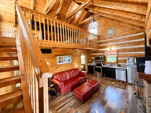 A wooden cabin interior with a loft bedroom, a red leather sofa, a kitchen area, and a dining table. - Blu's Blessed View 1 Bedroom Cabin (Sevierville)