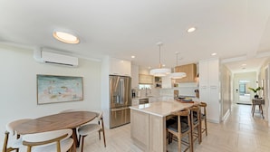 Una cocina moderna con una isla central, electrodomésticos de acero inoxidable, iluminación colgante y un comedor con mesa y sillas de madera.