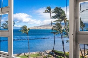 A view through a window of a coastal scene with palm trees, a clear sky, and a calm sea. - Menehune Shores, #507 2 Bedroom Condo (Kihei)