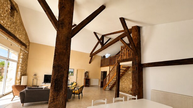 Un vaste espace de vie doté d’un haut plafond, de poutres apparentes, d’une table à manger avec des chaises, d’un canapé et d’un escalier menant à un niveau supérieur.