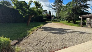 Une allée de gravier mène à un bâtiment en pierre surmonté d’un toit en tuiles, qui est entouré de verdure et d’arbres.