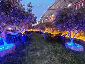 An outdoor seating area with tables and chairs, surrounded by trees and illuminated by string lights.