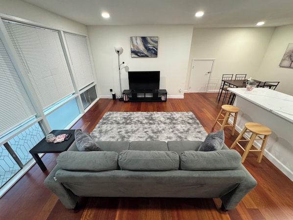 Ein modernes Wohnzimmer mit grauem Sofa, Kamin, Essbereich mit Barhocker und großem Fenster mit Jalousien.