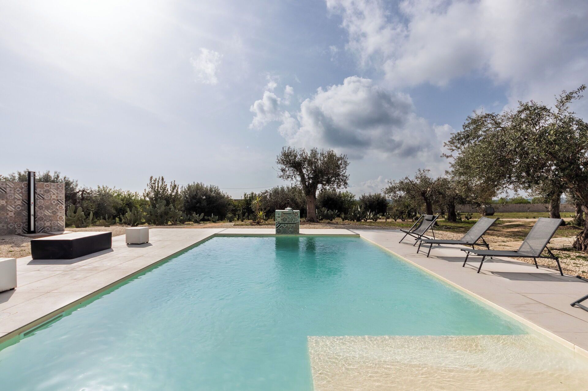 Une piscine rectangulaire à l’eau d’un bleu limpide, entourée d’une terrasse dallée. On y trouve des chaises longues et une petite structure blanche.