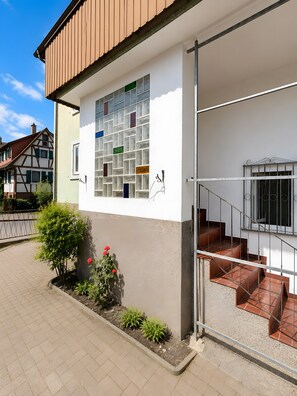 Ein Gebäude mit weißer Fassade, einem Fenster mit bunten Glassteinen, einer Treppe mit Metallgeländern und einem kleinen Garten mit Pflanzen und Blumen.