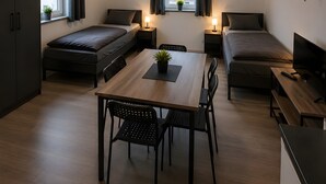 Ein kleines Zimmer mit Bett, Schreibtisch und Stuhl, ein kleiner Tisch mit Pflanze, einem Fernseher und einem Fenster mit Vorhängen.