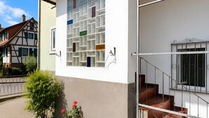 Ein Gebäude mit weißer Fassade, einem Fenster mit bunten Glassteinen, einer Treppe mit Metallgeländern und einem kleinen Garten mit Pflanzen und Blumen.