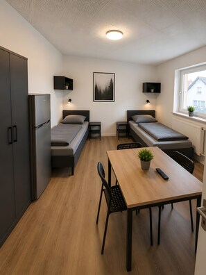 Une chambre compacte comprenant un coin cuisine, deux lits simples, une table à manger avec des chaises, une plante en pot et une fenêtre avec vue.