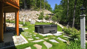 Un chemin de pierres menant à une terrasse en bois, entourée d’un jardin luxuriant et d’une colline rocheuse.