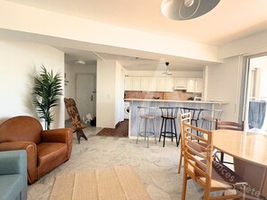 A modern kitchen with white cabinets, a central island with stools, and a dining area with a wooden table and chairs. - 011 / GRAND T3 RESIDENCE LE FRANCE (Sète)