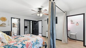 Een slaapkamer met een bed, een plafondventilator, een kledingkast, een badkamer en een klein zithoekje.
