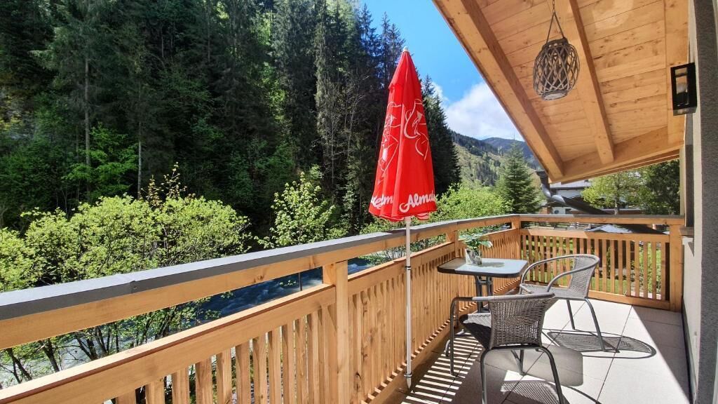 A balcony with a wooden railing, a red umbrella, and a table with chairs.