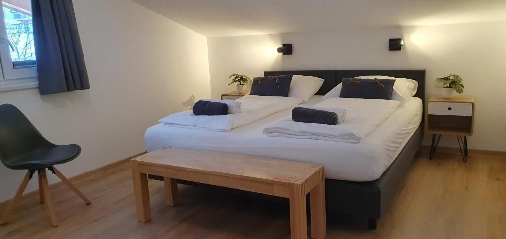 Cama doble con sábanas blancas, mesita de noche, una silla y una pequeña mesa en una habitación luminosa con piso de madera.