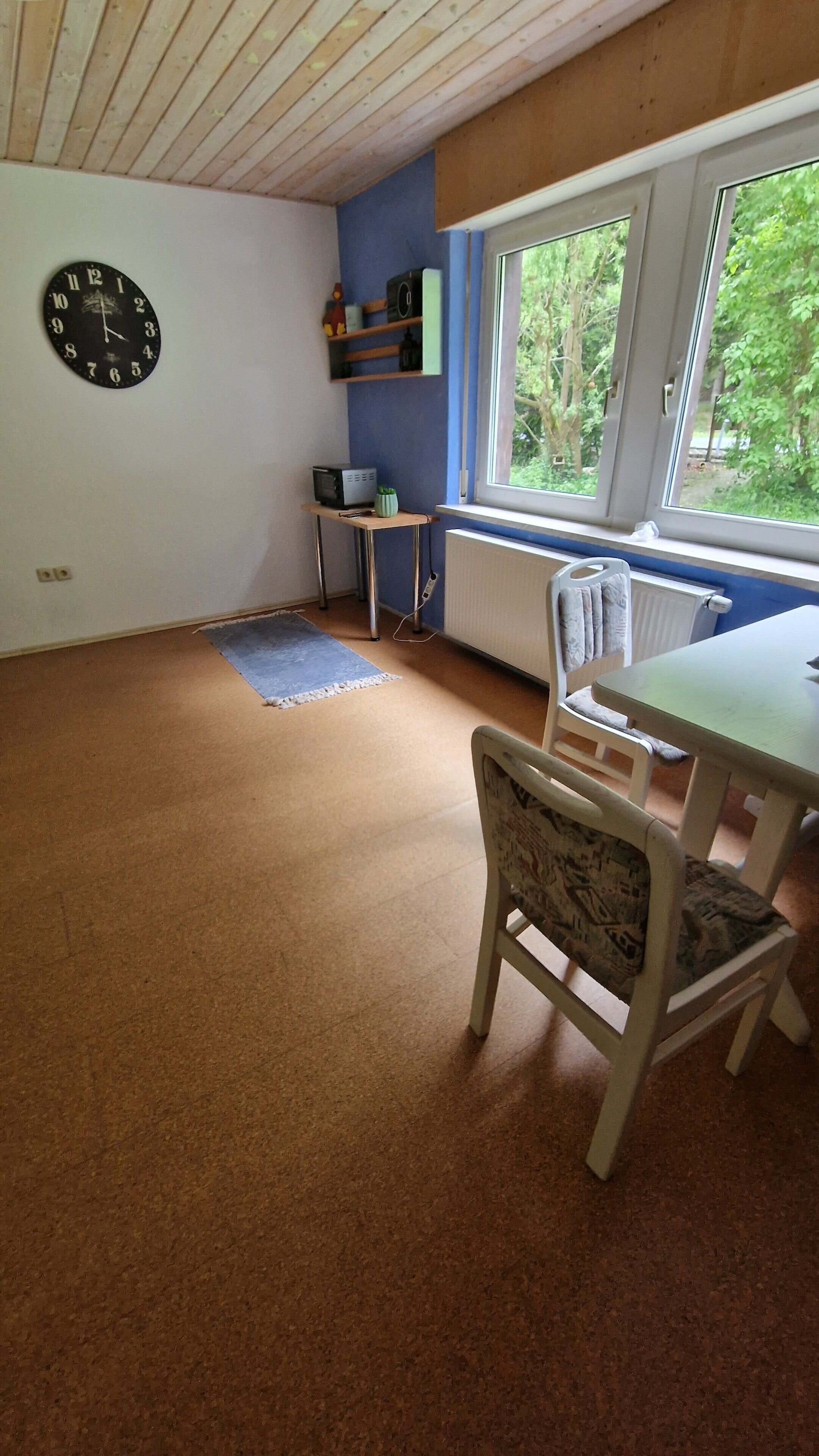 Ein Zimmer mit Holzboden, einer Wanduhr, einem Fenster mit Blick auf Bäume, einem Tisch mit Stühlen und einem Heizkörper.
