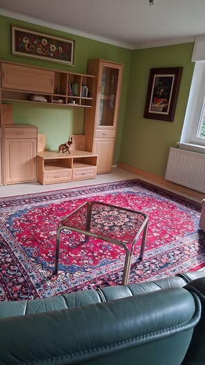 Ein Zimmer mit einem gemusterten Teppich, einem Holzkasten, einem Bücherregal und einem gerahmten Bild an der Wand.