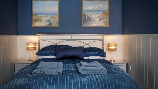 Un dormitorio con una cama, dos cuadros colgados en la pared, lámparas de noche y una cama bien hecha con toallas dobladas.